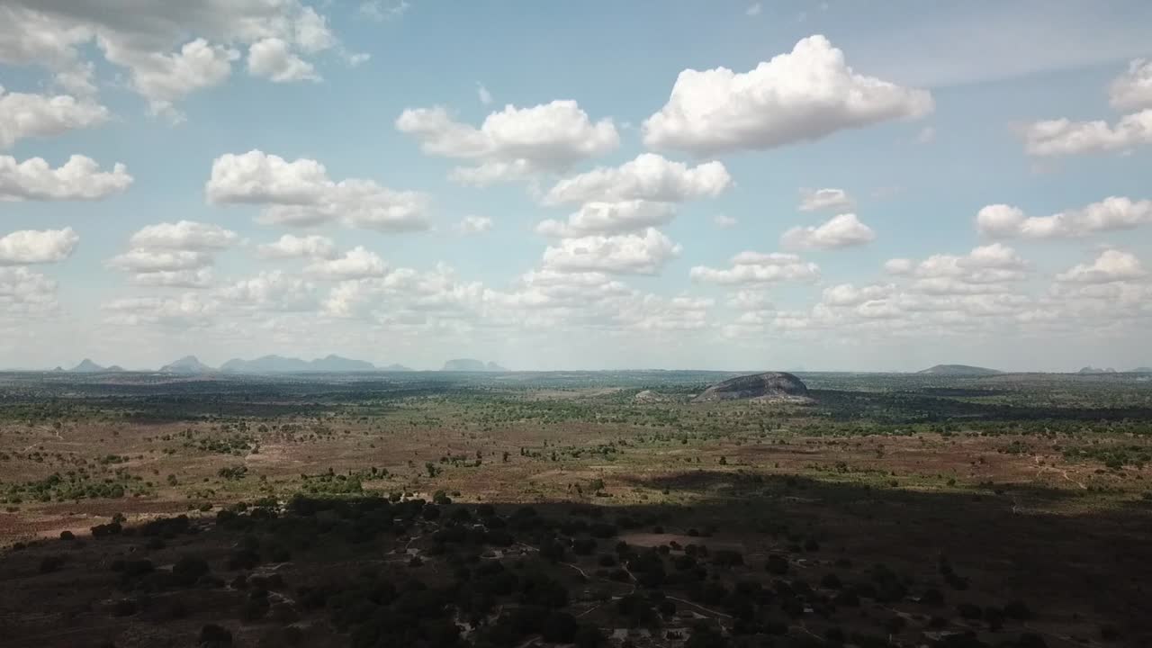 Wide aerial landscape of northern Mozambique (Nampula region), with dry savanna, woodland stretching to the horizon and isolated inselberg hills rising from the plain beneath scattered cumulus clouds