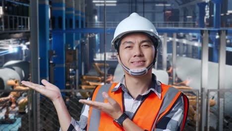 close up of asian male engineer with safety helmet standing in factory manufacture of wind turbines. smiling and pointing to side recommends about something while robotic arm working