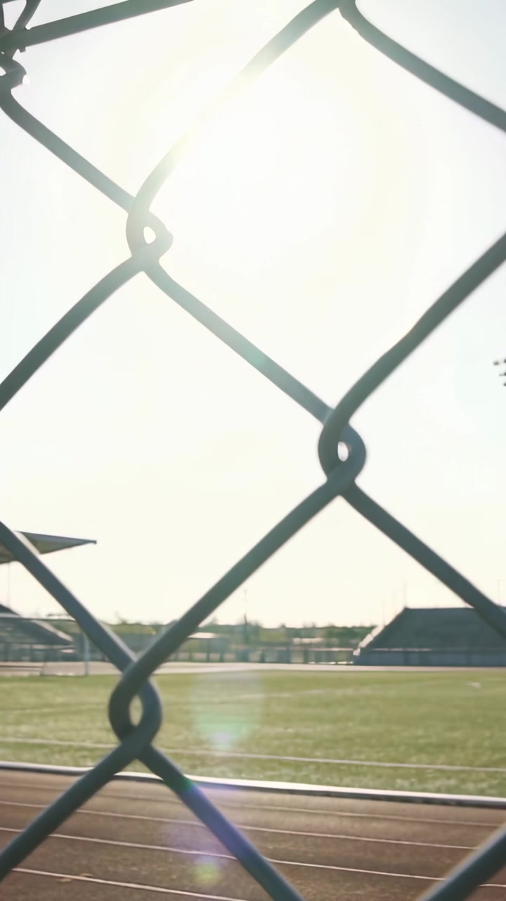 Vertical video: Camera panning, chain-link fence framing sun while revealing field at track, flare