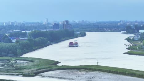 Vista-Aérea-Panorámica-De-Un-Barco-De-Contenedores-Cargado-Navegando-En-El-Río-Noord-Bajo-Un-Cielo-Gris-Nebuloso-Con-Paisaje-Urbano-En-El-Fondo,-Los-Países-Bajos