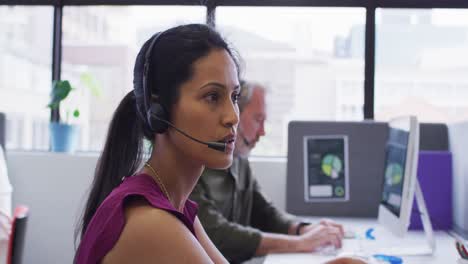 Mixed-race-businesswoman-sitting-using-computer-talking-on-phone-headset-in-office