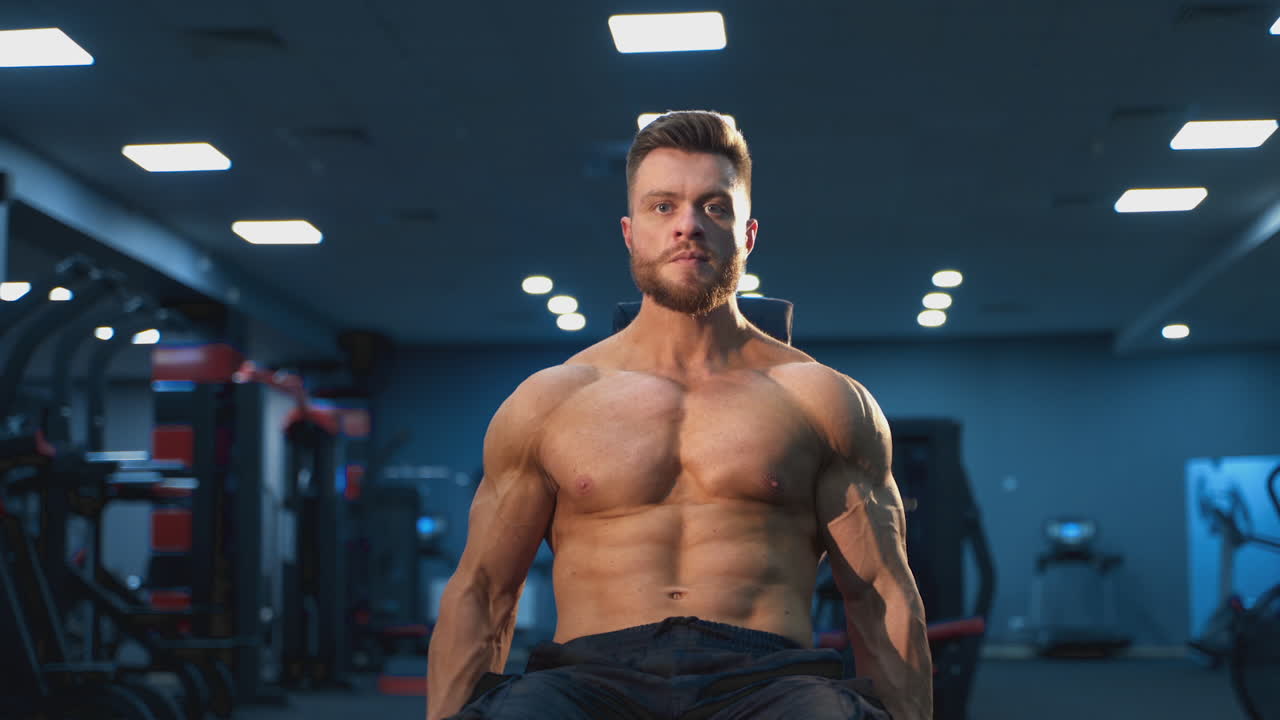 Concentrated young athlete doing lifting exercises. Portrait of a handsome bearded sportsman training with dumbbells in the gym. Workout of a bodybuilder.