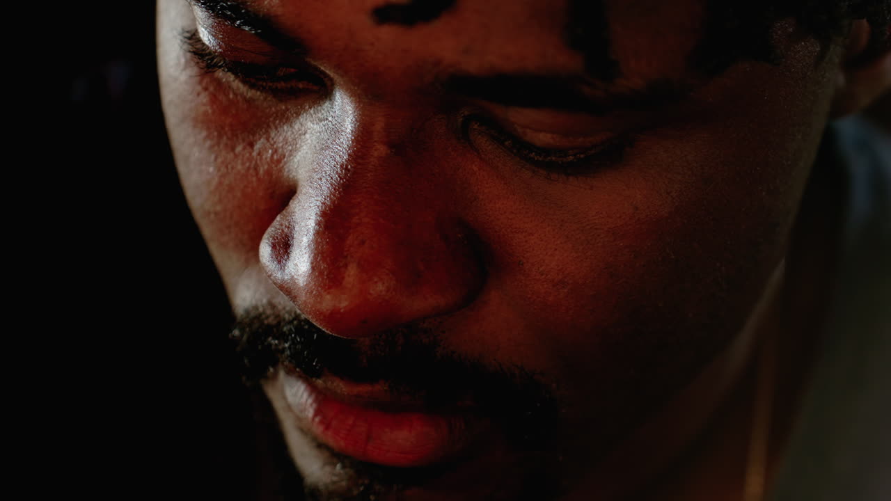 Close up of dark skin man with focused expression looking downward, showing intense concentration and thoughtful demeanor in soft natural light with blurred background