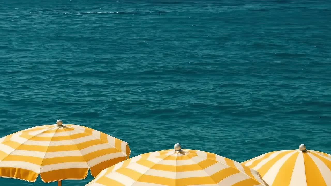 Video dall'alto di ombrelloni a strisce gialle in riva al mare, che cattura un'atmosfera da spiaggia serena.