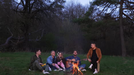 few friends having fun in the nature, playing guitar, singing, frying marshmallows, laughing. cozy summer evening picnic by the fire.