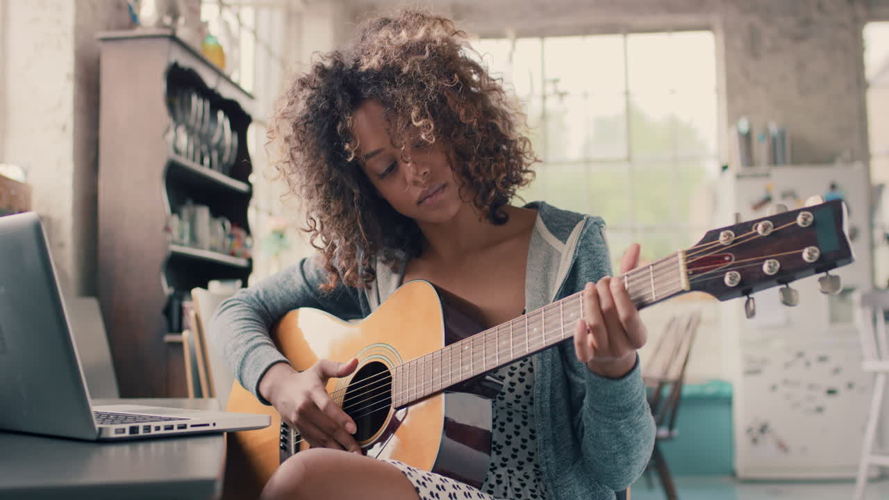 chica joven de raza mixta atractiva aprendiendo a tocar la guitarra usando una computadora portátil