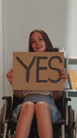 cheerful woman with disability turns poster with word yes to camera with smile on face. young person with muscular dystrophy sits in wheelchair demanding tolerance