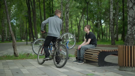 young boy with his head bowed, seated beside an upside-down bicycle with spinning tires, is approached by another boy riding a bicycle, the two begin to converse in a park setting