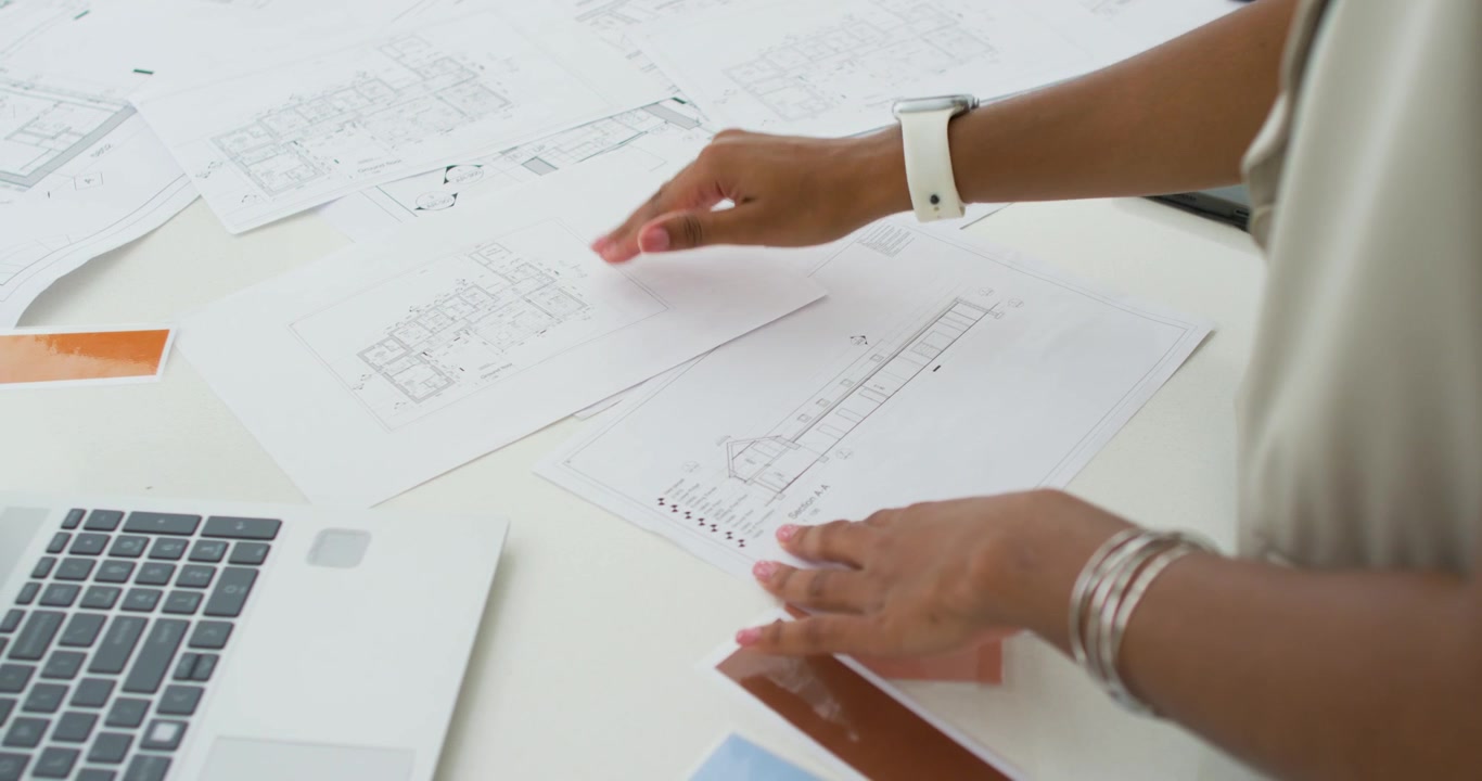 Adult female smoothing and aligning printed design plans at desk for review with laptop and tablet