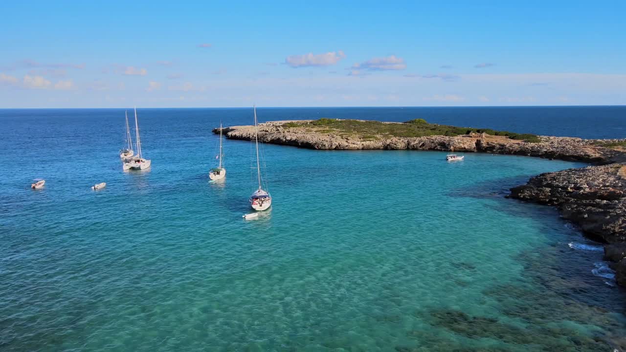 Breathtaking view of several sailing boats anchored in the vibrant turquoise waters of Cala Varques. The sun shines brightly over the stunning coastline of Mallorca, creating a serene atmosphere.