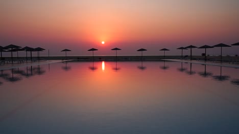 perfect red sunset view from tropical hotel resort lounge with infinity pool, beach umbrellas and their reflection