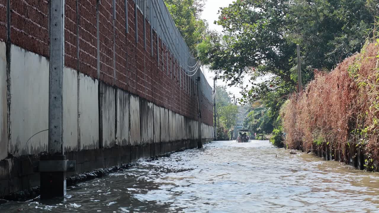 A boat navigates a narrow, polluted canal in Bangkok, Thailand. The scene captures murky water, overgrown vegetation, and urban surroundings