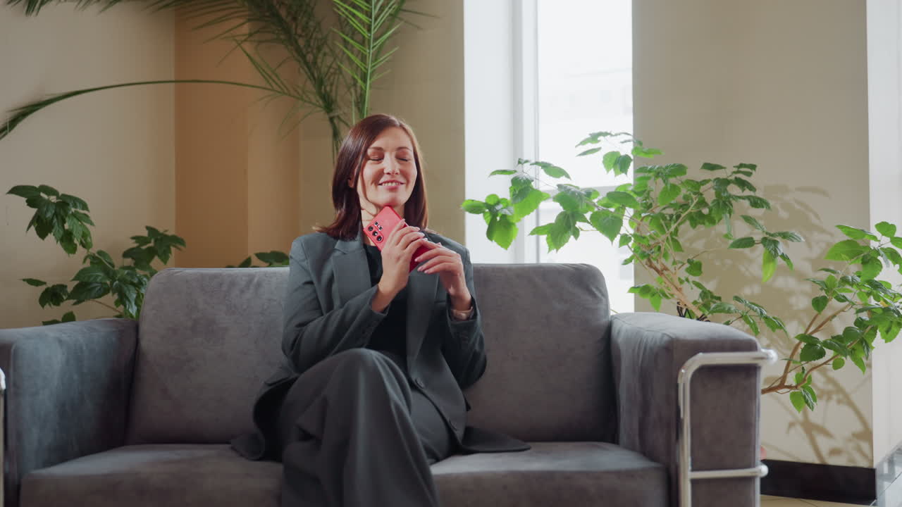 Businesswoman in suit sitting on modern sofa holding smartphone with thoughtful smile, relaxing in bright office lounge surrounded by green indoor plants and natural light from large window