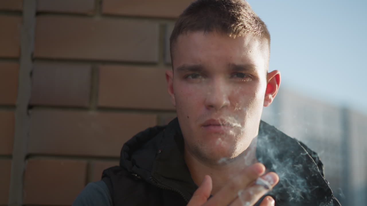 student with serious expression holds cigarette between fingers and exhales smoke slowly while leaning on brick wall, with blurry modern building in background and warm sunlight on face