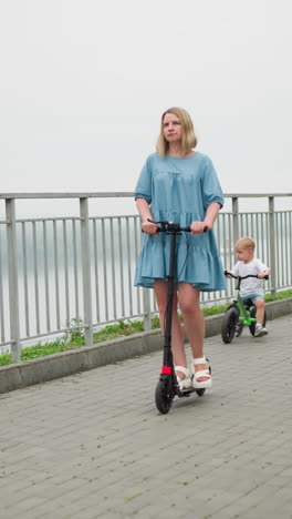 a woman rides a scooter with a thoughtful expression, followed by a young boy on a bicycle, they move along a bridge, with a scenic water body in the blurred background
