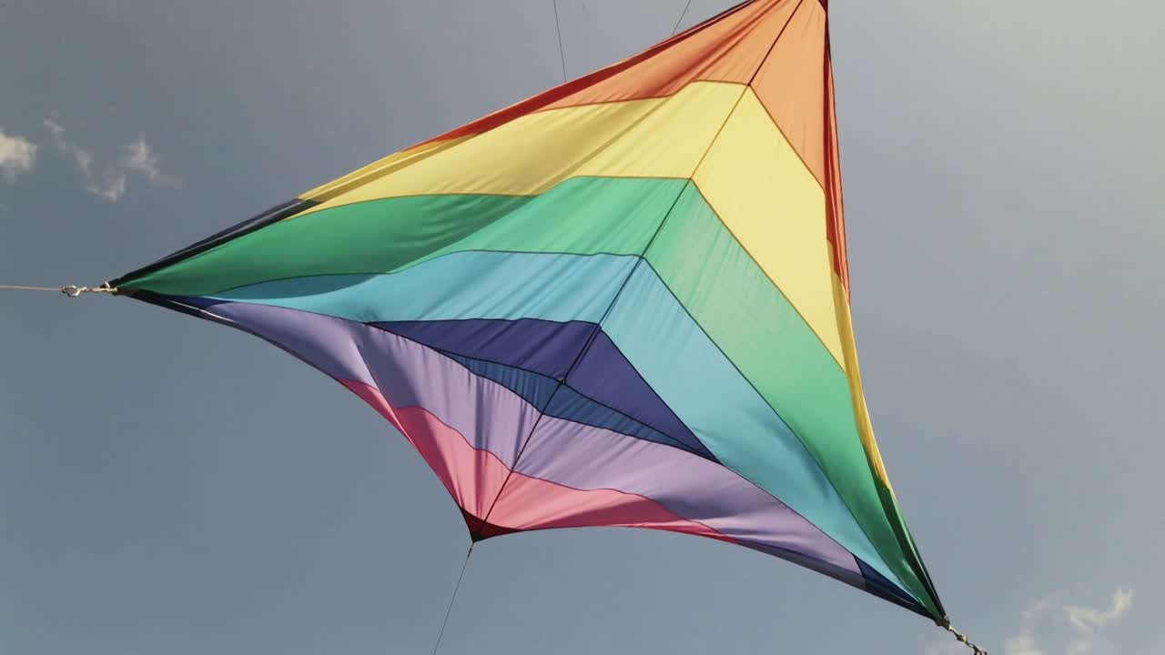 Aquilone delta triangolare arcobaleno che scivola e si gonfia con la brezza sopra un parco, con briglie e cavi di ancoraggio visibili.