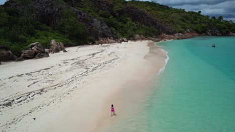 woman in red bathing suit walking down sandy tropical beach alone on curieuse island the seychelles with boat anchored off shore