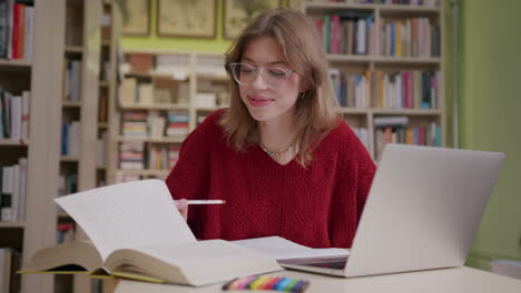 niña sonriente con anteojos estudia con libro abierto y portátil en la biblioteca