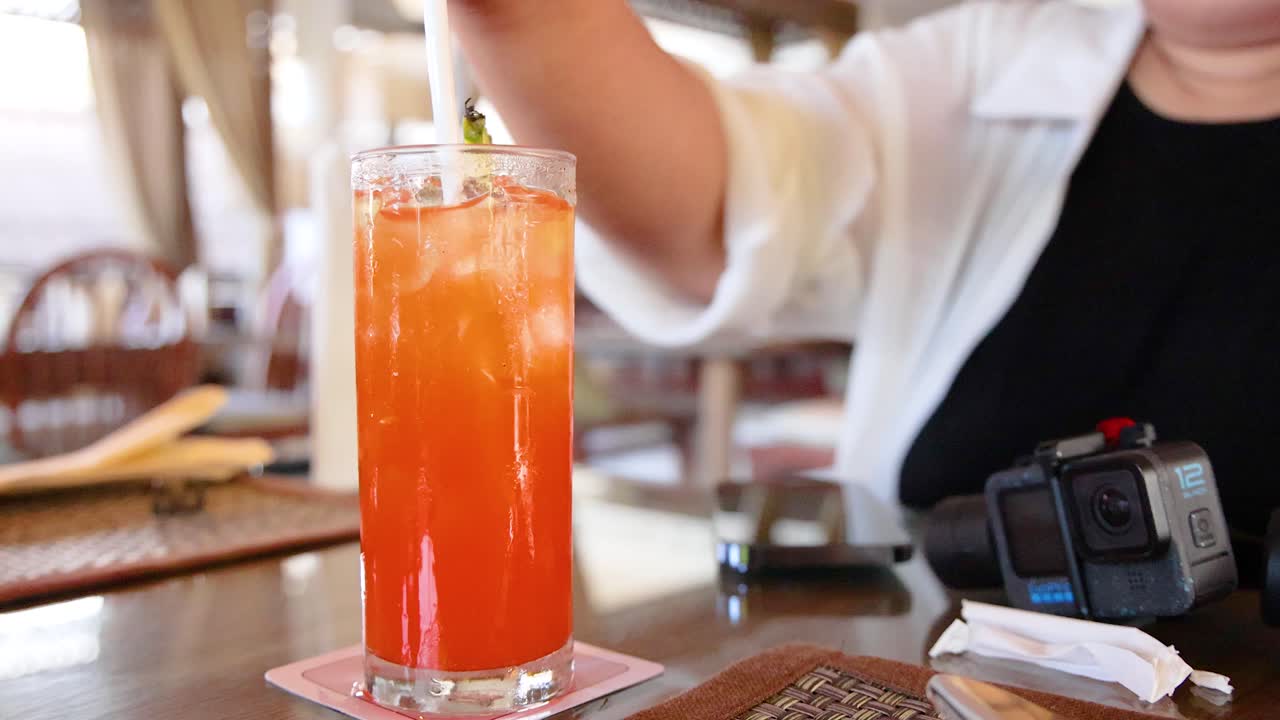 A vibrant orange cocktail is stirred by a person in a relaxed café environment, captured with natural lighting