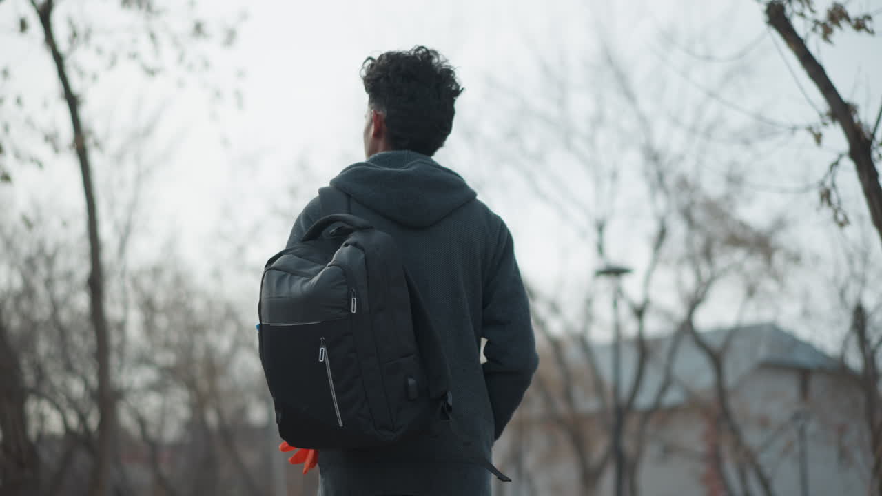 Man with backpack standing outdoors facing away, holding orange gloves in hand, wearing dark hoodie and jeans, surrounded by bare winter trees, peaceful park environment, contemplative and solitary mood
