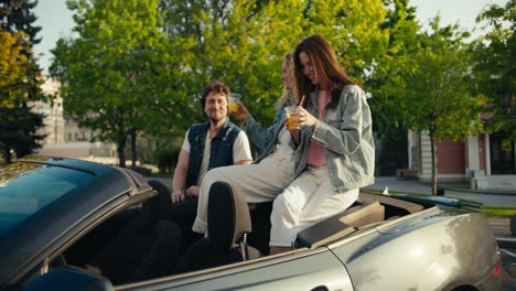 Group-of-happy-young-friends-in-denim-jackets-sitting-in-cabriolet-and-drinking-yellow-cocktails.-A-brunette-and-a-blonde-in