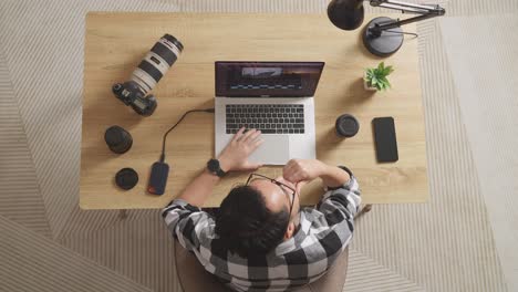 top view of asian male editor thinking about the footage then raising index finger while sitting in the workspace using a laptop next to the camera editing the video at home