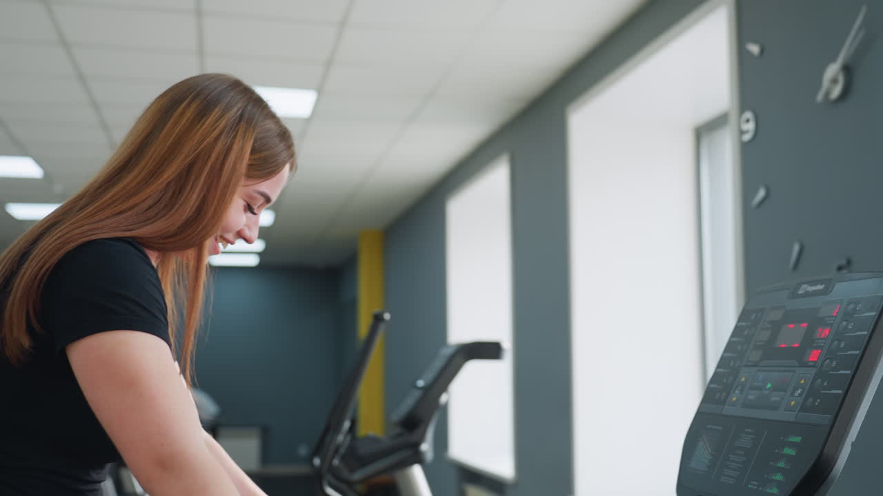 lady with long hair smiling gently while stepping off treadmill after workout in brightly lit gym with modern interior, visible treadmill control panel and large wall clock