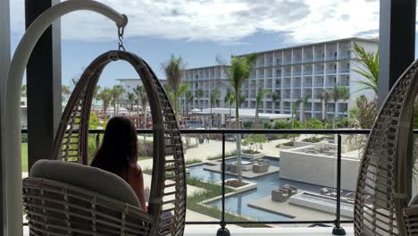 Young-woman,-swinging-in-hanging-chair,-hammock,-overlooking-pool-area-in-luxury-all-inclusive-resort-vacation,-female-harmony-and-lifestyle,-meditation,-mindfulness,-tranquility