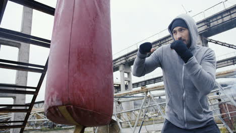 Bottom-view-of-caucasian-man-in-grey-hoodie-hitting-a-punching-bag-outdoors-an-abandoned-factory-on-a-cloudy-morning