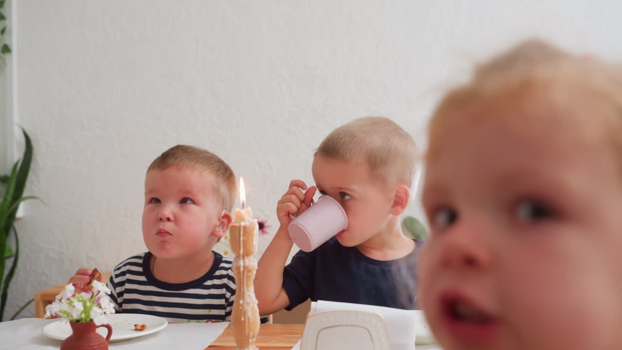 Group of young kids sitting at table during kindergarten snack time, one drinking from pink cup while another eats beside candlelight, girl in foreground looking curiously toward camera