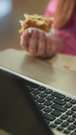 vista de la mano de una mujer trabajando en su portátil con una hamburguesa parcialmente comida en su mano izquierda, haciendo una pausa para tomar su bebida de la mesa, colocándola de nuevo, y continuando su trabajo, el fondo ligeramente borroso