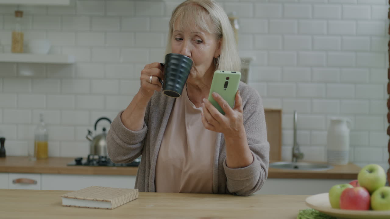 Senior Woman Using Phone and Drinking Coffee in Kitchen
