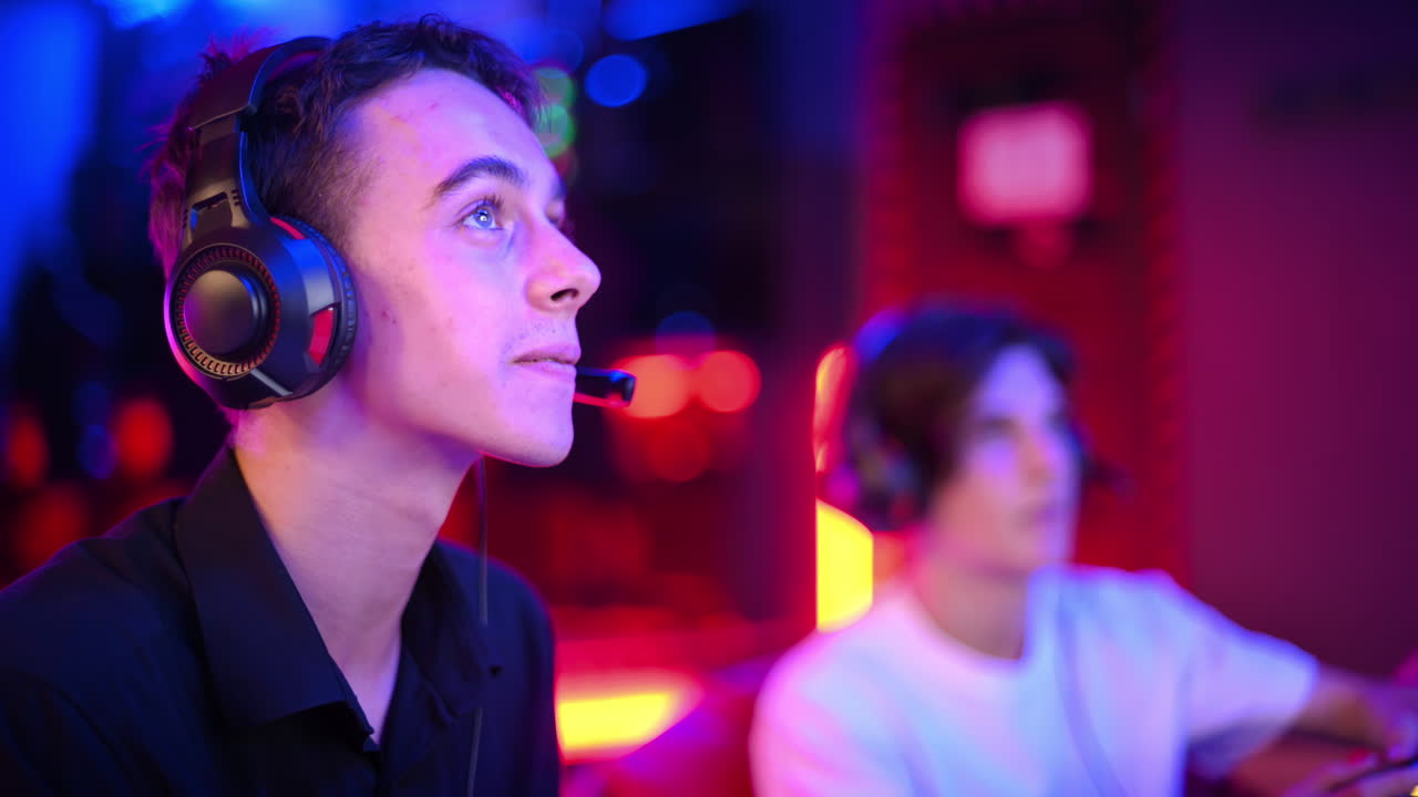 Two teen friends in headphones are talking and playing a game console using gamepads, talking and laughing while sitting on bean bags. Blue and red illumination