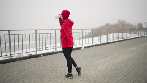 lady in red hoodie and beanie stands outdoors in serene winter park setting with snow-covered ground, opens pink water bottle, sips water, near iron railing, trees, and river in misty background