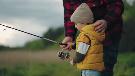 curious little boy is learning to catch fish by rod in lake grandfather is teaching grandson