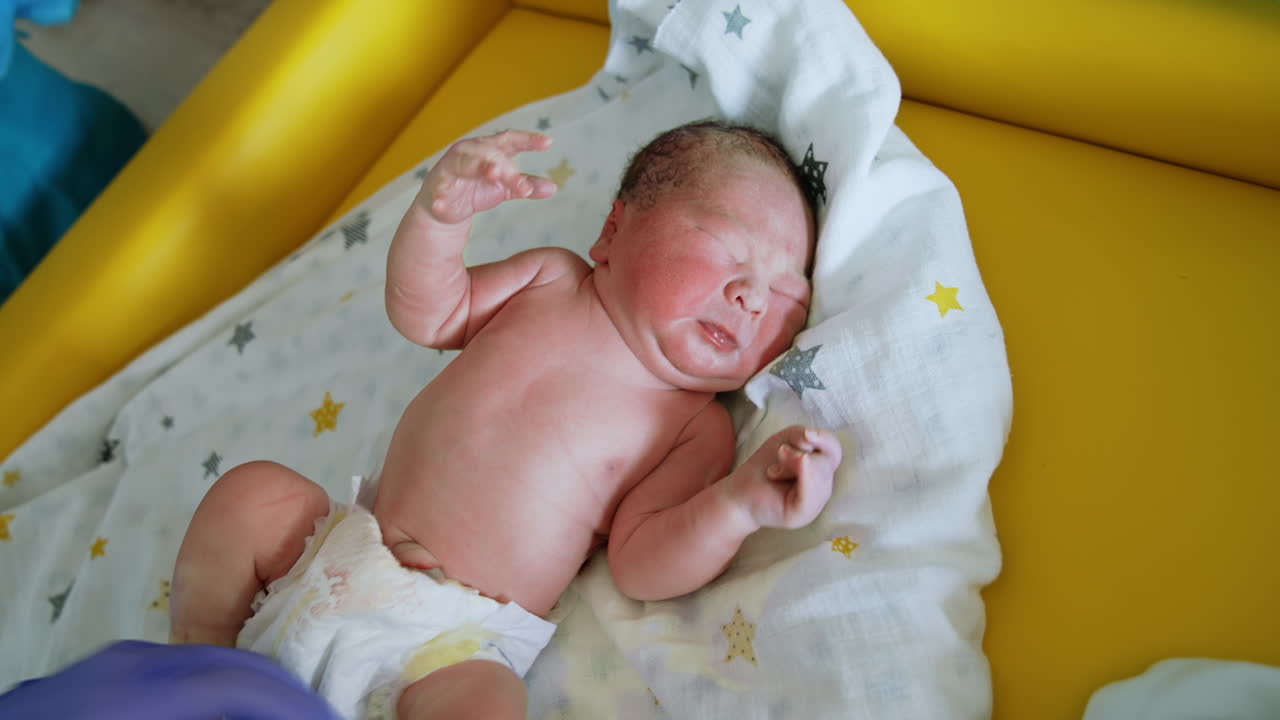 Newborn baby is crying while nurse takes care of him. Child is unhappy to be undressed. Top view.