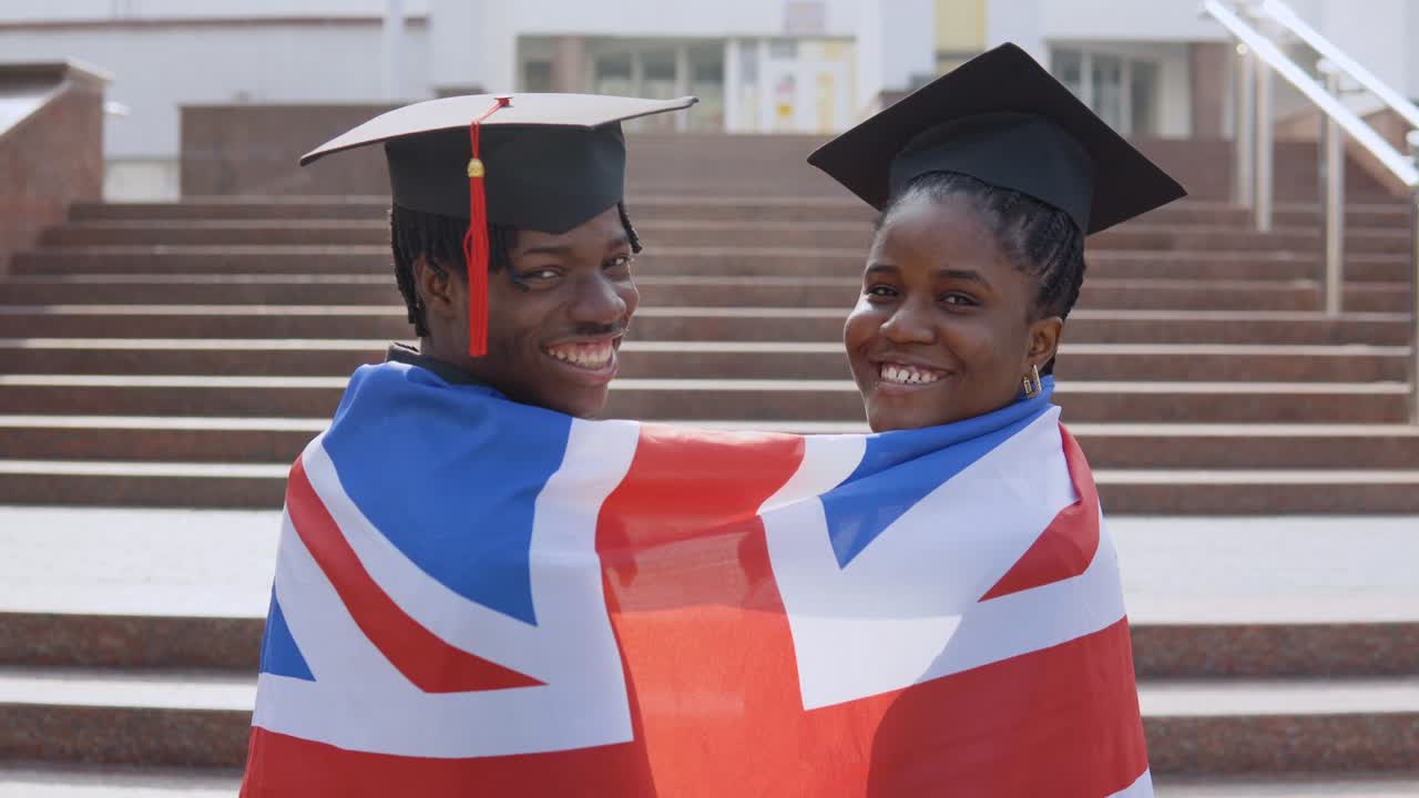 hombre y mujer afroamericanos de pie uno al lado del otro con las espaldas hacia la cámara en túnicas negras y sombreros cuadrados de estudiantes graduados con la bandera británica en los hombros. mirando a la cámara