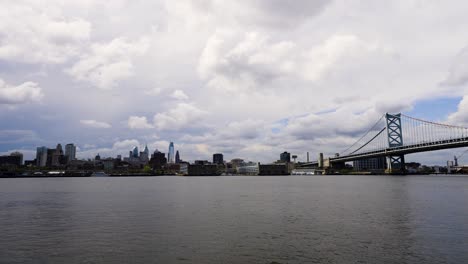 Skyline-Von-Philadelphia-über-Dem-Ruhigen-Delaware-River-Mit-Der-Benjamin-Franklin-Bridge-An-Einem-Wolkigen,-Warmen-Tag-Tief-Und-Weit-Blau