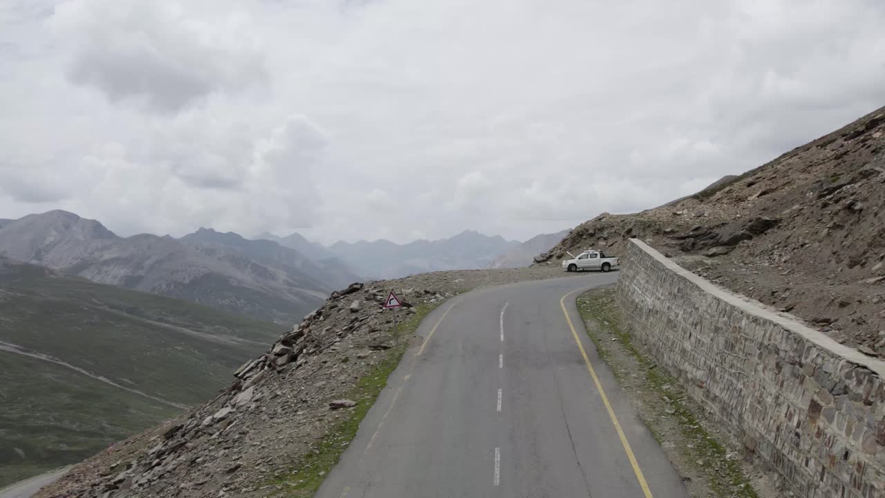 Curved mountain road with vehicles overlooking vast green valley and rugged peaks at Babusar Pass. Khyber Pakhtunkhwa, Pakistan