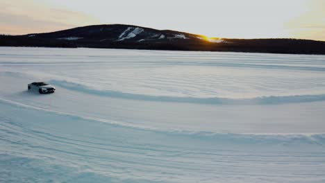 Drohnenaufnahme-Eines-Sportwagens,-Der-Bei-Sonnenuntergang-Auf-Einem-Zugefrorenen-See-Rutscht,-Mit-Sonnenuntergang-Und-Bergen-Im-Hintergrund-Im-Polarkreis