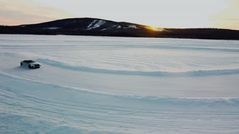 toma de drone de un auto deportivo deslizándose sobre un lago congelado durante la puesta de sol con puesta de sol y montañas al fondo en el círculo ártico
