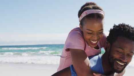 Pareja-Afroamericana-Tomando-Selfie-Con-Teléfono-Inteligente,-Hombre-Llevando-A-Una-Mujer-A-Cuestas-En-La-Playa