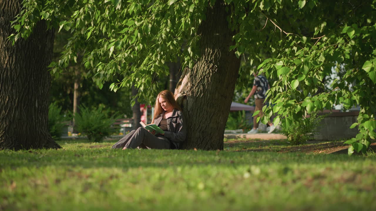 야외에 앉아있는 여성, 잔디에 있는 나무에 기대어, 따뜻한 빛 아래 책을 읽고, 나무 잎이 바람에 부드럽게 흔들리고, 배경에는 건물과 는 여성이 약간 흐릿하게 보입니다.