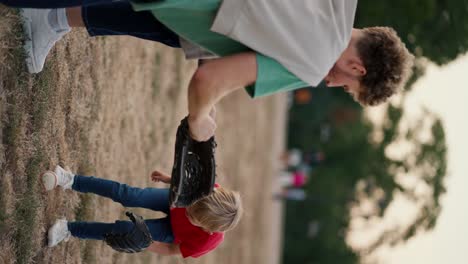 Vertical-video:-A-little-blond-boy-in-a-red-T-shirt-with-a-basketball-glove-throws-a-basketball-into-the-basketball-glove-of-his-dad-in-a-Green-T-shirt-on-the-court-in-the-park