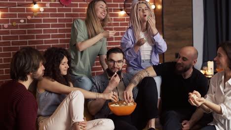 friends grabbing popcorn from a bowl while sitting on a sofa at home