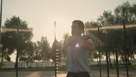 concentrated sportsman doing deltoid and tricep stretch exercises in outdoor court at sunset