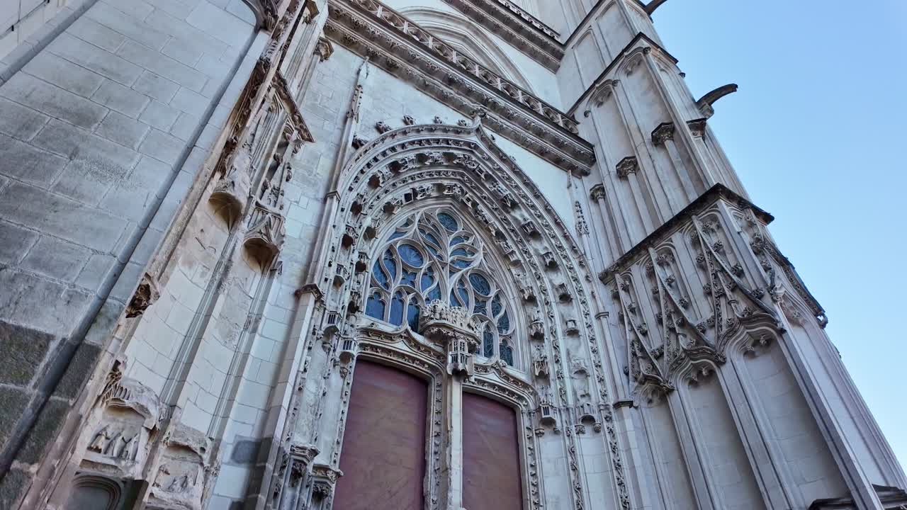 Movimiento lento hacia adelante en la entrada de la Catedral de Nantes, seguido de una suave inclinación hacia arriba, revelando arcos góticos, columnas esculpidas, sillería ornamentada y cielo azul arriba.