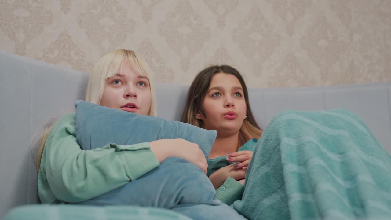 two young girls wrapped in blankets sit on bed watching scary movie scene with tense expressions, one clutching pillow tightly while other clasps hands anxiously showing visible fear and anticipation