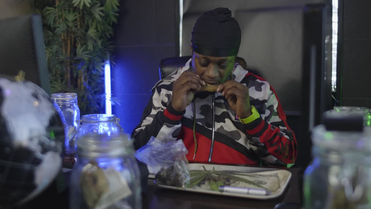 straight on view of a black man licking a blunt to seal to tobacco wrapper around the weed there is a rolling tray on the desk and a jar of money in the foreground