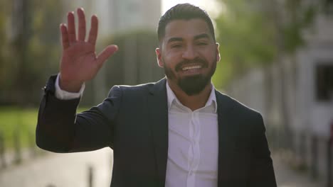 uomo d'affari felice che agita la mano e guarda la telecamera all'aperto